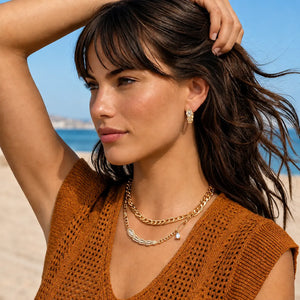 A woman on a beach wearing layered gold and silver necklaces, a gold hoop earring, and a brown knit top.