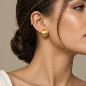 A close-up of a woman's ear wearing a textured gold stud earring and a smaller gold ear cuff, against a soft beige background.