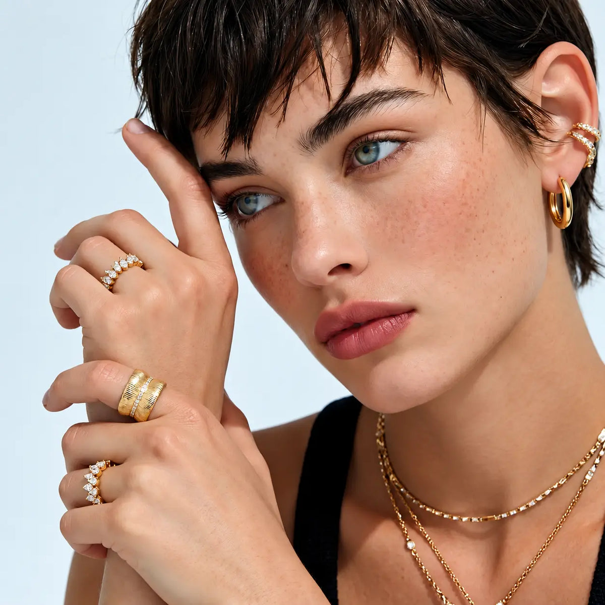 Close-up of a model wearing multiple gold rings with clear stones, including a stacked band ring, a ring with a single stone, and a delicate chain necklace, against a light blue background.