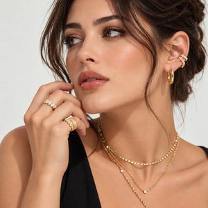 A close-up lifestyle shot of a woman wearing gold jewelry, including layered gold necklaces, rings, and hoop earrings, against a clean light gray background.