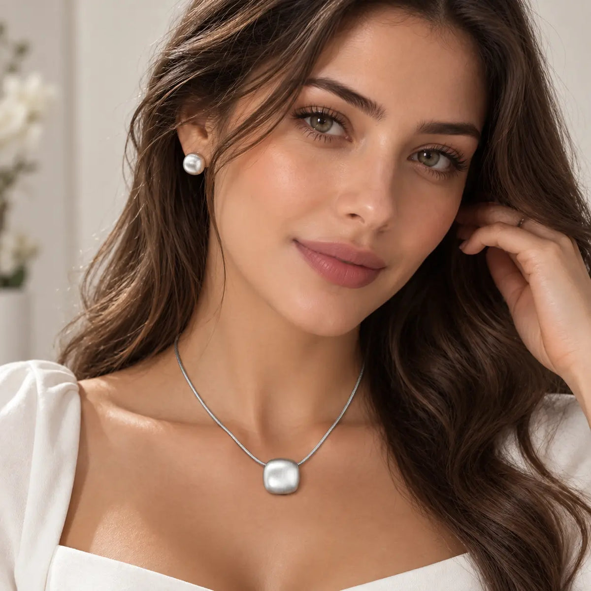 A woman with long wavy brown hair models a silver pendant necklace with a rounded square charm and a matching silver stud earring against a soft, light background.