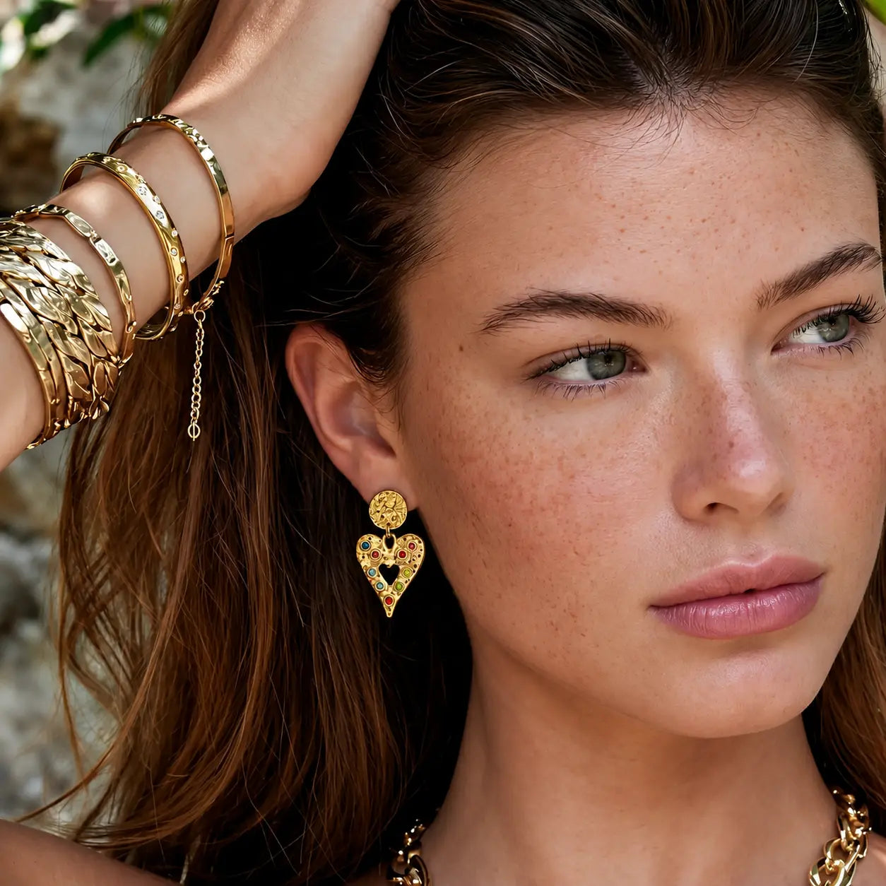 A close-up lifestyle shot of a woman with freckles wearing a gold heart-shaped dangle earring with a small red heart in the center, along with stacked gold bangles on her wrist.
