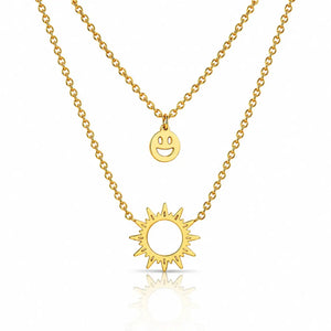 A double-layered gold necklace set featuring a delicate cable chain with a small smiley face disc pendant and a thicker link chain with a sunburst pendant, displayed on a clean white background with a soft reflection.