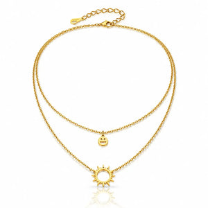 A double-layered gold necklace featuring a delicate chain with a small smiley face charm and a larger sun pendant, displayed on a clean white background with a subtle reflection.