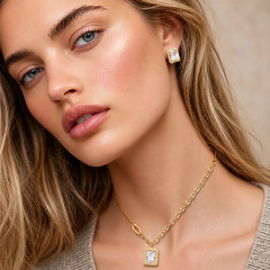 Close-up of a woman wearing a gold chain necklace with a square pendant and a matching stud earring, both featuring clear gemstones in a gold setting.