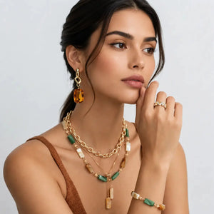 A woman models a coordinated jewelry set featuring a gold-tone layered necklace with green and cream beads, matching drop earrings, and rings, set against a clean light gray background.