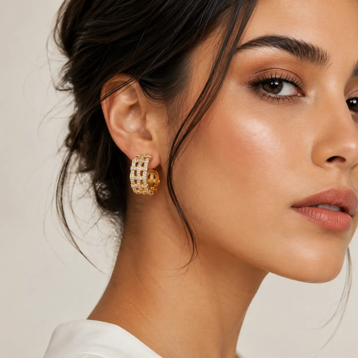 A close-up of a woman's ear wearing a gold hoop earring with a textured, openwork pattern and a small gold ball accent at the back, against a soft beige background.