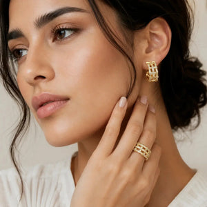 A close-up lifestyle shot of a woman wearing a gold hoop earring and a matching gold ring with small clear stones, set against a soft beige background.