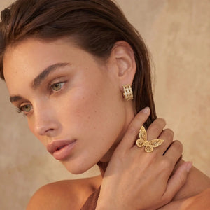 Woman wearing gold butterfly earrings and ring against a beige background