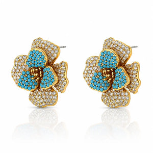 A pair of gold flower-shaped stud earrings embellished with turquoise blue stones and clear pavé crystals, shown on a white background with a soft reflection below.