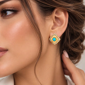 A close-up of a woman's ear wearing a gold-toned geometric stud earring with a bright turquoise center and small clear stones around it.