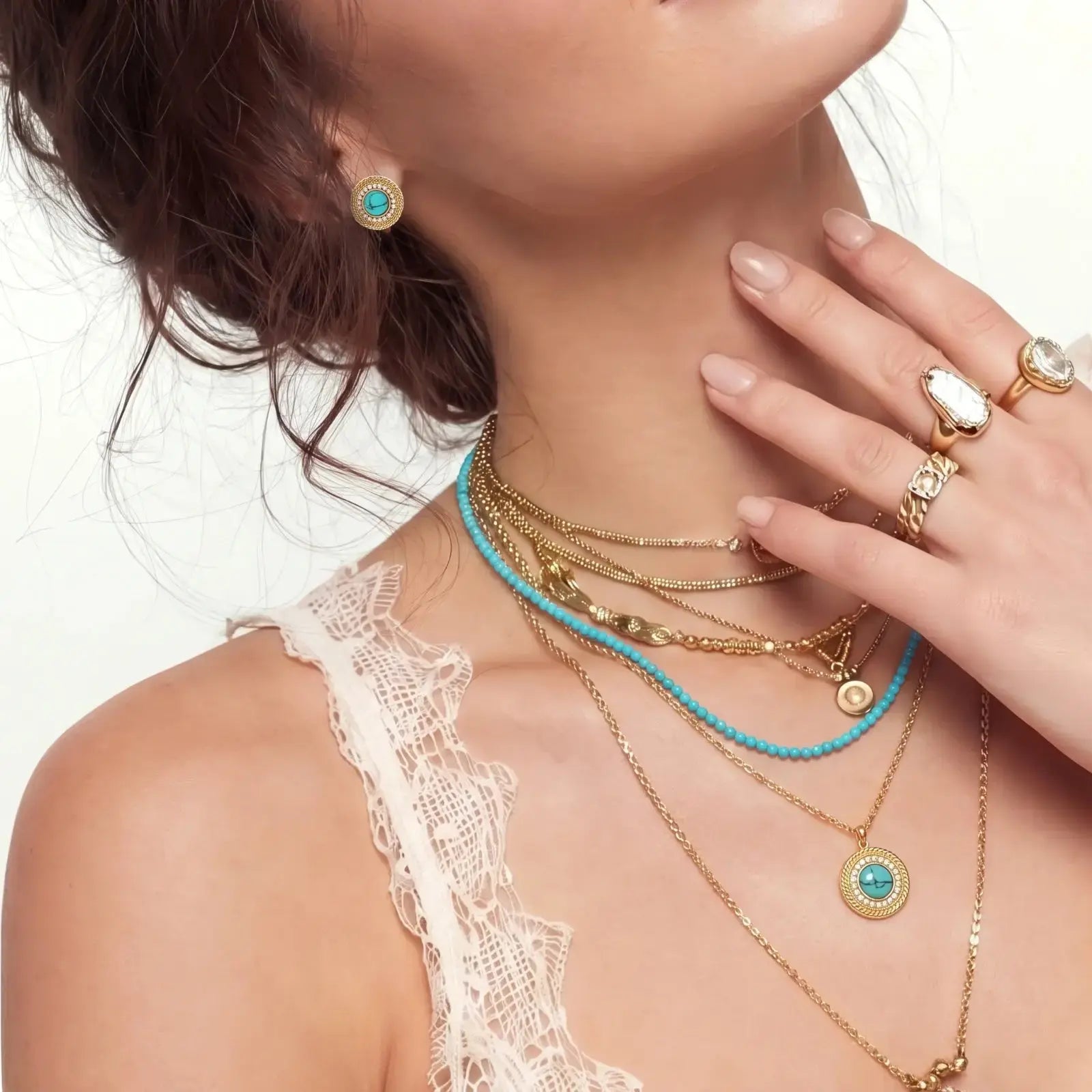 Woman wearing gold necklaces, turquoise earrings, and rings on a neutral background
