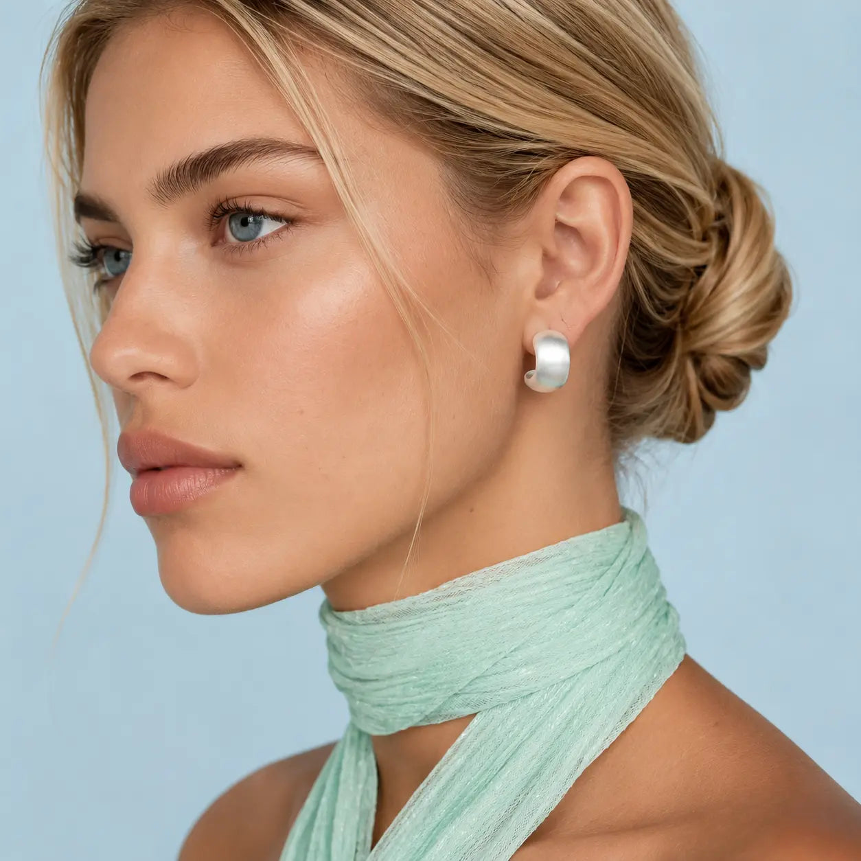 A close-up side profile of a woman with blonde hair in a low bun, wearing a small, chunky white pearl stud earring. She is wearing a mint green halter top against a solid light blue background.