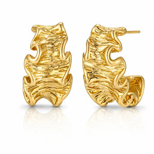 A pair of gold huggie hoop earrings with an organic, wavy, textured surface, displayed on a clean white background with a soft reflection below.