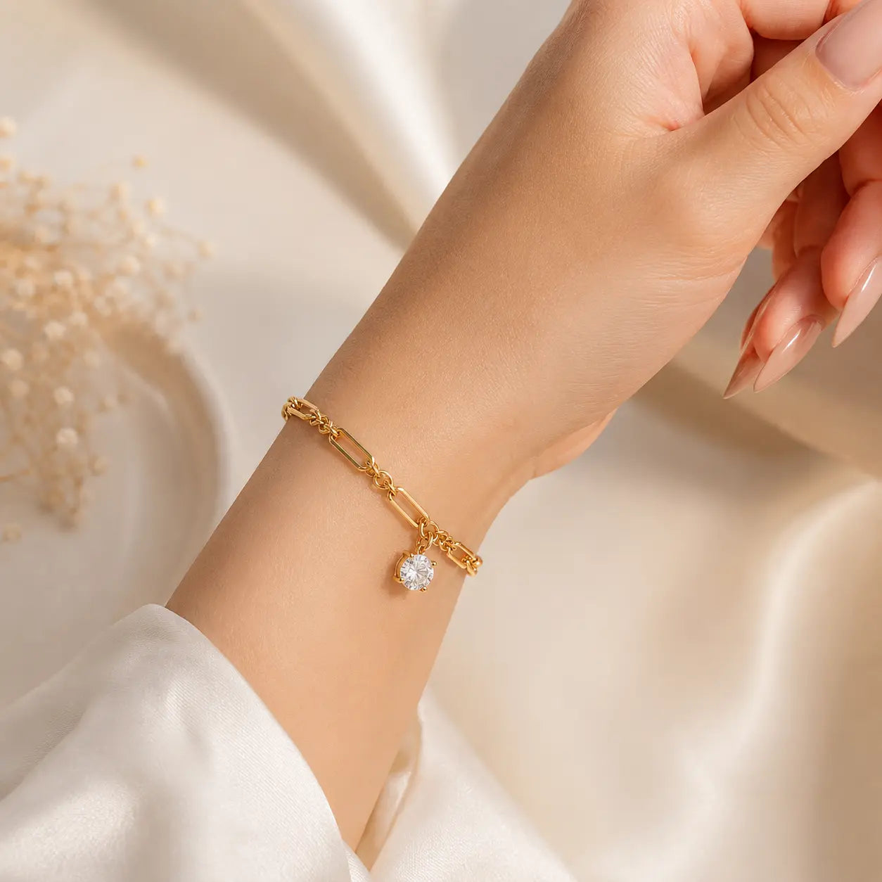 A gold-toned multi-strand bracelet with three heart-shaped crystal charms, shown worn on a woman's wrist. The hand has manicured nails and is posed against a warm, softly lit cream background with decorative dried flowers in the corner.