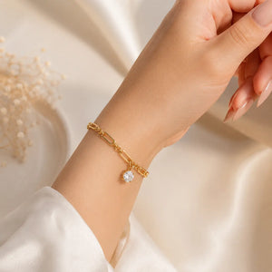 A gold-toned multi-strand bracelet with three heart-shaped crystal charms, shown worn on a woman's wrist. The hand has manicured nails and is posed against a warm, softly lit cream background with decorative dried flowers in the corner.