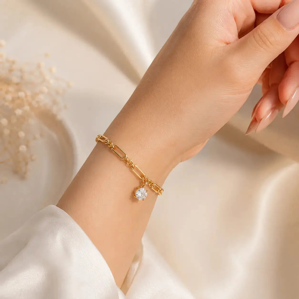 A gold-toned multi-strand bracelet with three heart-shaped crystal charms, shown worn on a woman's wrist. The hand has manicured nails and is posed against a warm, softly lit cream background with decorative dried flowers in the corner.