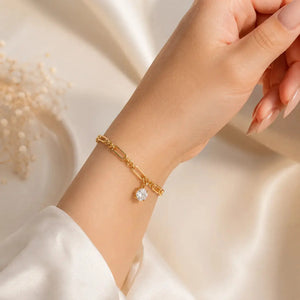 A gold-toned multi-strand bracelet with three heart-shaped crystal charms, shown worn on a woman's wrist. The hand has manicured nails and is posed against a warm, softly lit cream background with decorative dried flowers in the corner.