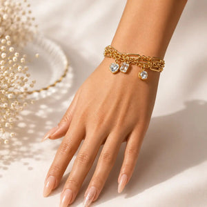 A gold-toned multi-strand bracelet with three heart-shaped crystal charms, shown worn on a woman's wrist. The hand has manicured nails and is posed against a warm, softly lit cream background with decorative dried flowers in the corner.