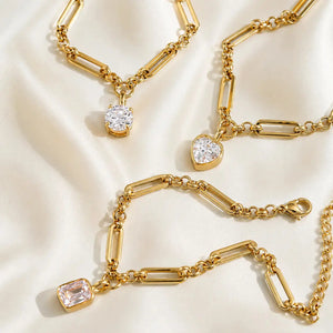 A gold paperclip chain bracelet with a rectangular clear gemstone pendant, displayed on a clean white background with a subtle reflection.