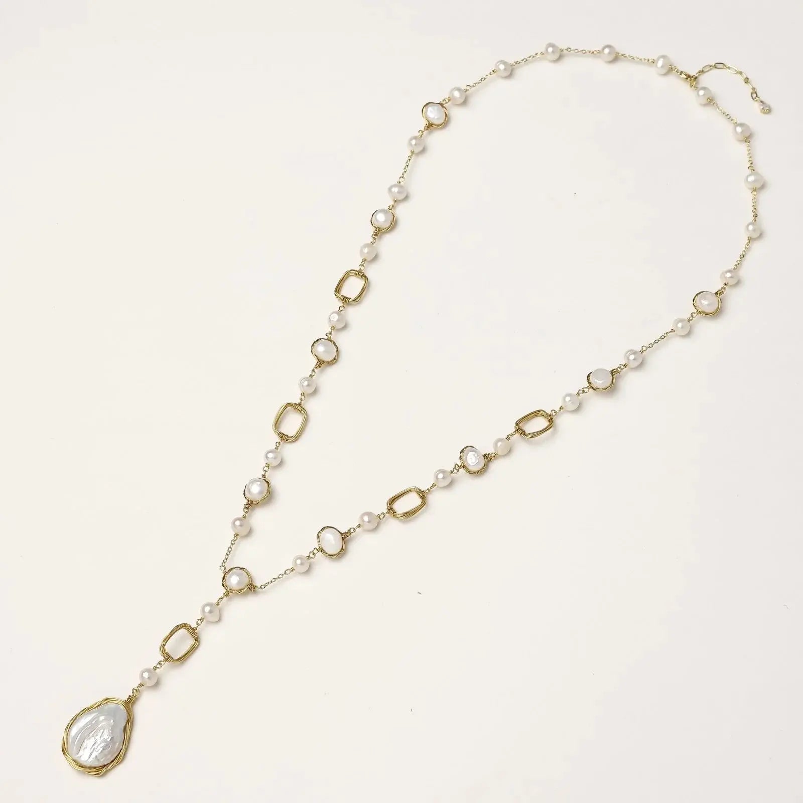 Y-shaped gold necklace with alternating geometric links, freshwater pearls, and baroque pearl pendant on cream background
