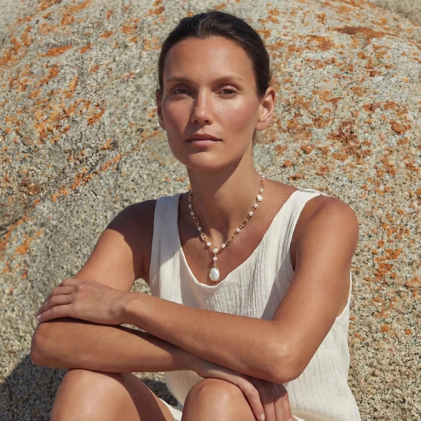 Woman sitting against a textured stone wall wearing a white sleeveless top and necklace.
