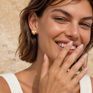 A close-up lifestyle shot of a woman with short brown hair smiling, wearing a small gold heart stud earring and a gold ring on her finger, set against a warm beige textured background.