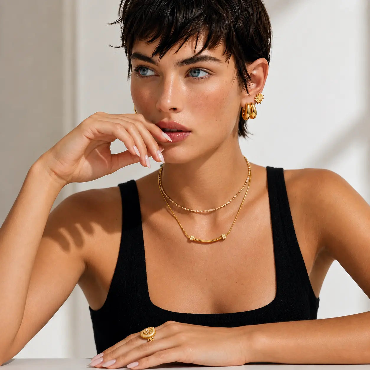 A woman with short dark hair wearing layered gold necklaces, hoop earrings, and a ring, posing in a black ribbed tank top against a bright white background.