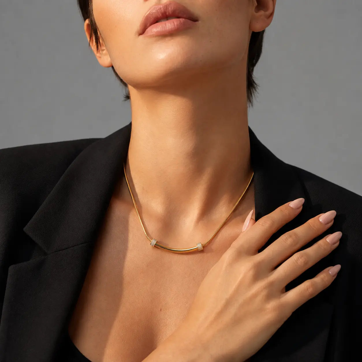 A close-up of a model wearing a delicate gold necklace with small geometric clear stone accents, styled with a black blazer against a neutral gray background.