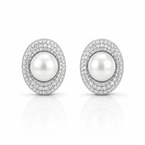 A pair of oval stud earrings featuring a central white pearl surrounded by a halo of small clear stones in a silver-toned setting, shown on a clean white background with a soft reflection below.