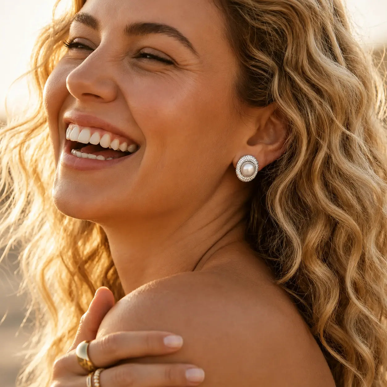A close-up lifestyle shot of a smiling woman with wavy blonde hair wearing a round silver stud earring with a white pearl center, set against a warm, sunlit outdoor background.