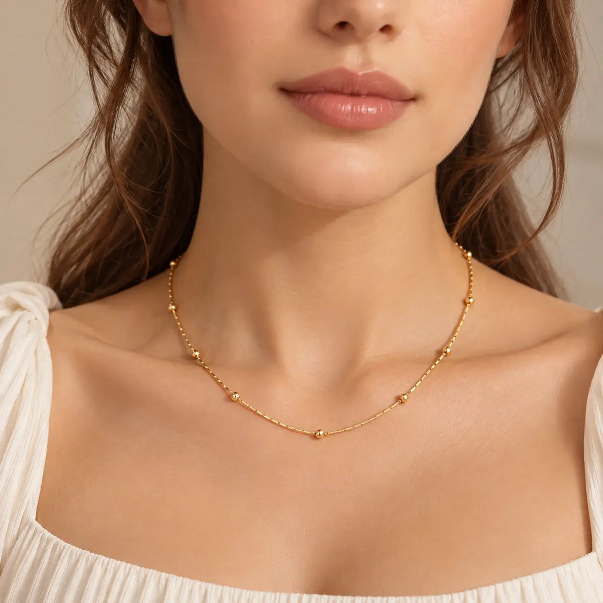A close-up of a woman wearing a delicate gold chain necklace with small bead details, styled with a white off-the-shoulder top against a soft beige background.