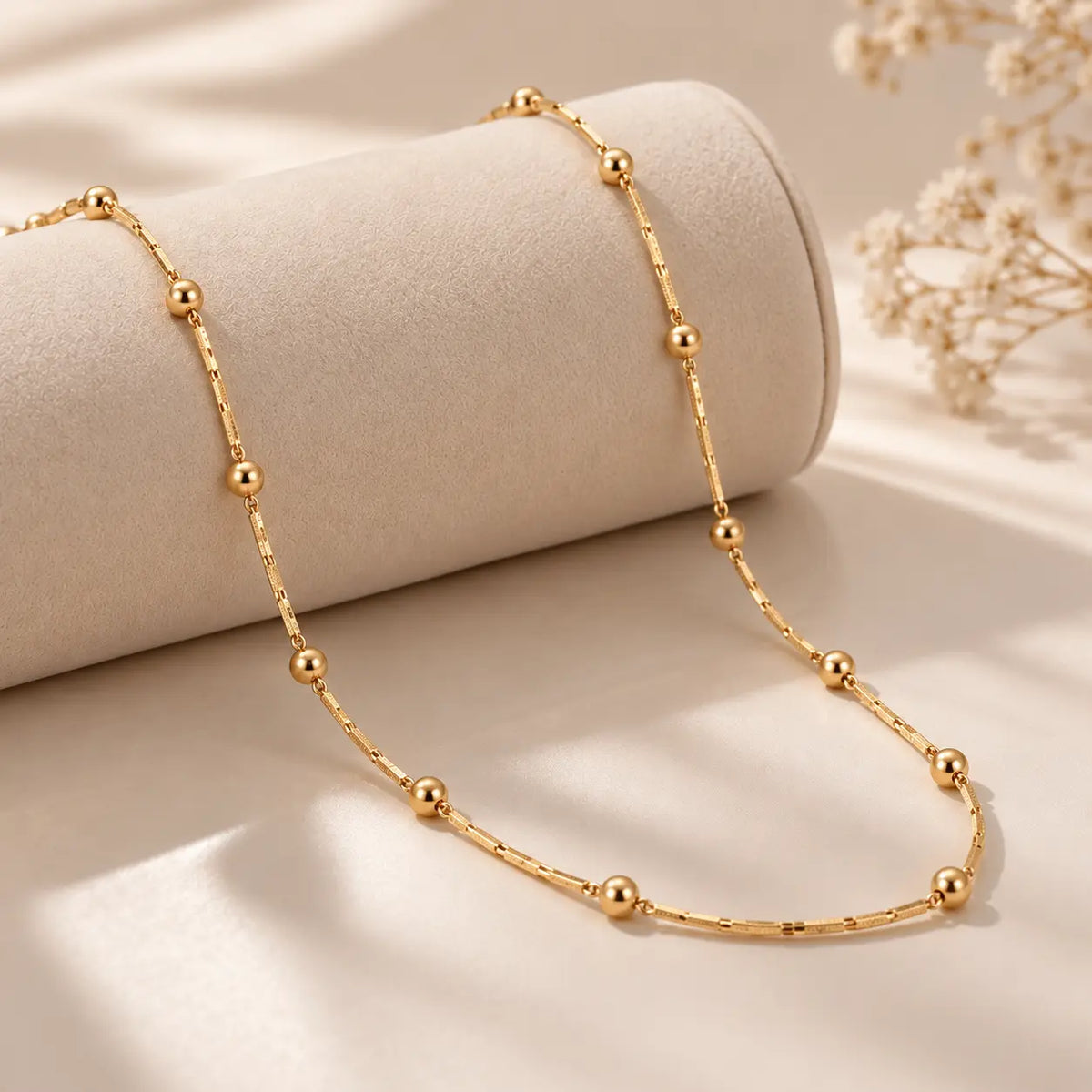 A delicate gold beaded chain necklace draped over a cylindrical display stand on a soft beige surface with dried floral decor in the background.