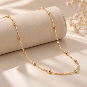 A delicate gold beaded chain necklace draped over a cylindrical display stand on a soft beige surface with dried floral decor in the background.