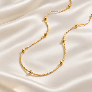 A delicate gold chain necklace with evenly spaced polished spherical beads, displayed on a draped cream satin fabric background.