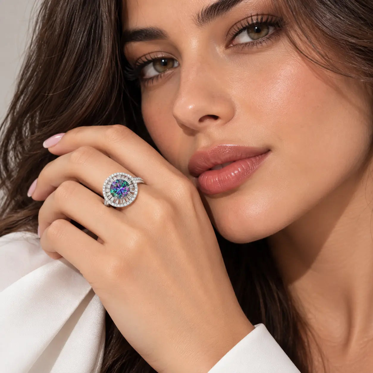 A close-up lifestyle shot of a woman wearing a silver-toned halo ring with a round center stone featuring blue and purple hues, set against a soft neutral background.