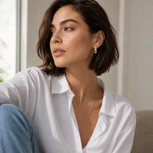 Woman wearing a white shirt and gold necklace, sitting indoors.