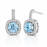 A pair of silver-toned drop earrings featuring a large round-cut light blue gemstone in a halo setting, with smaller clear stones along the top bar and around the square-shaped drop, shown on a white background with a soft reflection.