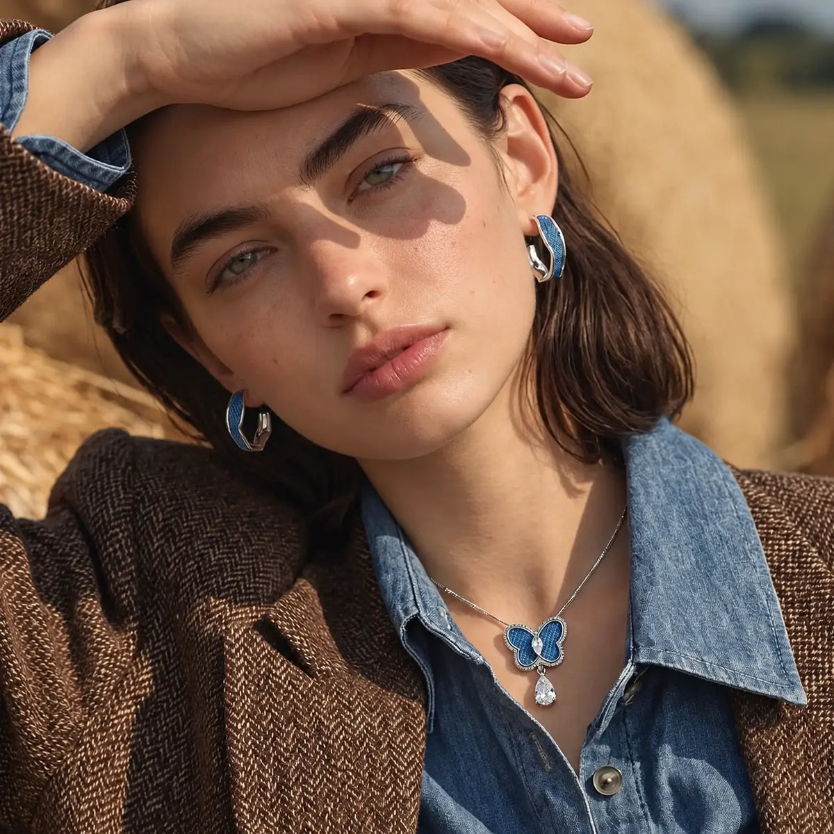 A woman wearing a silver necklace and matching stud earrings with blue butterfly details and a dangling clear crystal, styled with a denim strap top against 
