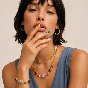 A close-up lifestyle shot of a model wearing a gold-toned jewelry set featuring a multi-colored stone necklace, matching bracelet, and hoop earrings. The model has short dark hair and is wearing a slate blue top against a plain beige background.
