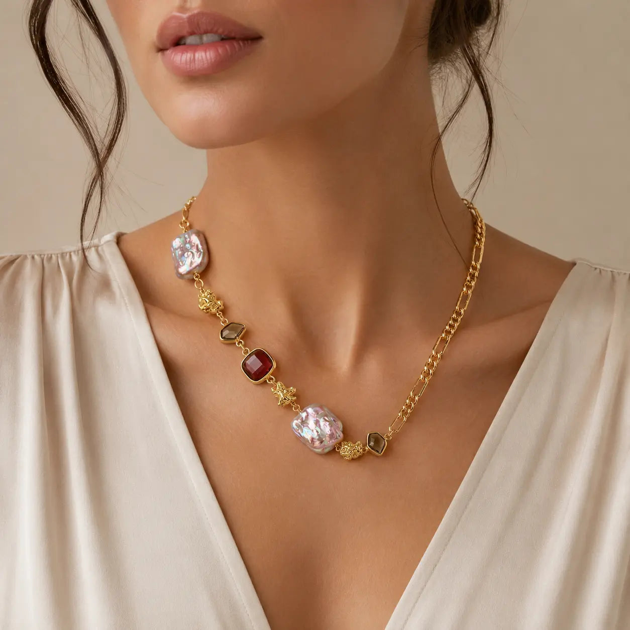 A gold chain necklace with irregular white and red gemstone accents, worn by a model in a cream-colored blouse against a warm beige background.