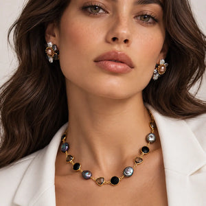 A close-up of a woman wearing a gold necklace with round and heart-shaped black and iridescent shell beads, matching shell stud earrings, and a white blazer.
