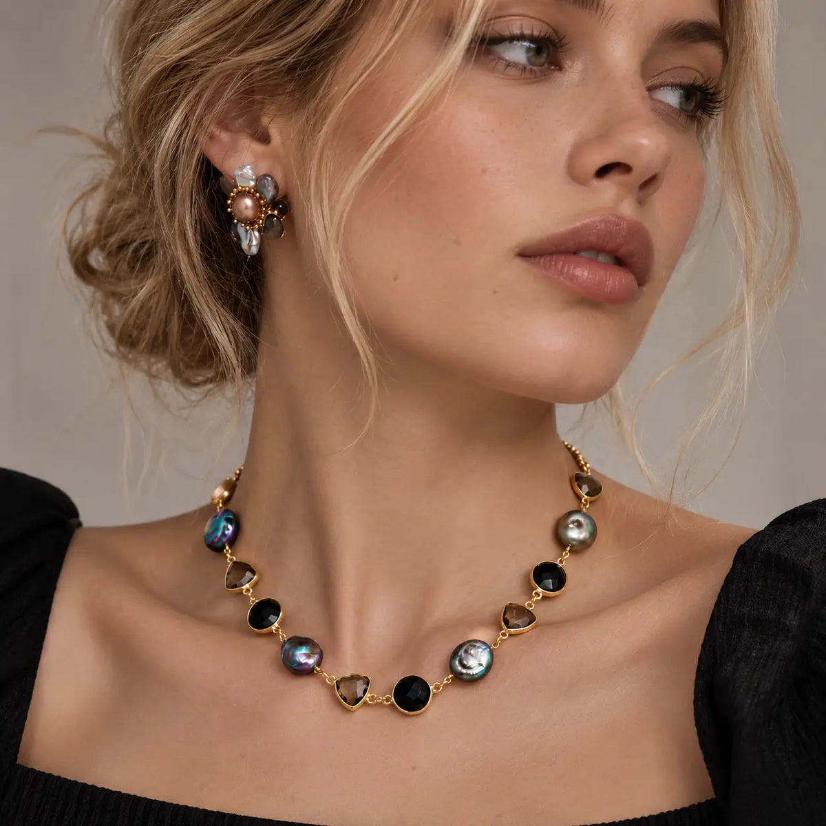 A close-up of a woman wearing a gold-toned necklace with oval iridescent shell-like stones and matching stud earrings against a soft beige background.