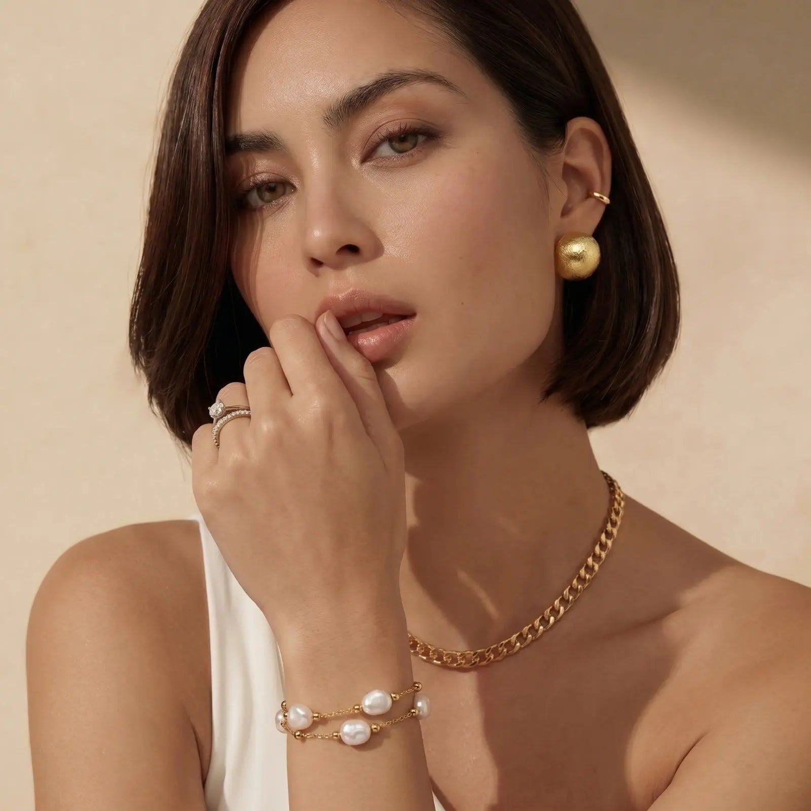 Woman wearing gold jewelry including earrings, necklace, and bracelet against a beige background
