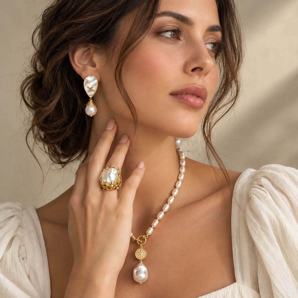 A close-up lifestyle shot of a woman wearing a matching gold and pearl jewelry set, including a necklace, drop earrings, and a ring, paired with a white blouse against a warm beige background.