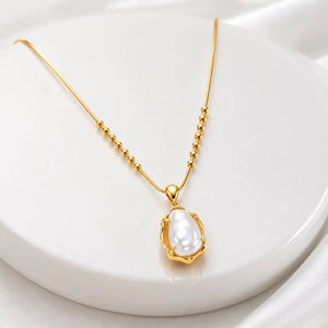 A gold chain necklace with a textured pendant featuring a white baroque pearl, displayed on a white circular pedestal.