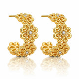 A pair of gold huggie hoop earrings with intricate floral filigree detailing and small clear crystal accents, displayed on a clean white background with a soft reflection below.
