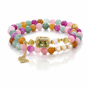 A multi-strand beaded bracelet featuring pastel pink, orange, green, and purple beads with white pearl-like accents, gold-toned metal details, and a central square green gemstone centerpiece, displayed on a clean white reflective surface.