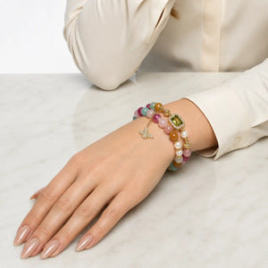 A close-up of a person's hand and wrist wearing a stack of colorful beaded bracelets with gold-toned charms, including a butterfly and a green gemstone accent, on a white marble surface.