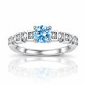 A silver-toned diamond ring featuring a round-cut light blue center stone and small clear accent stones set in square-shaped bands, shown on a white background with a soft reflection below.